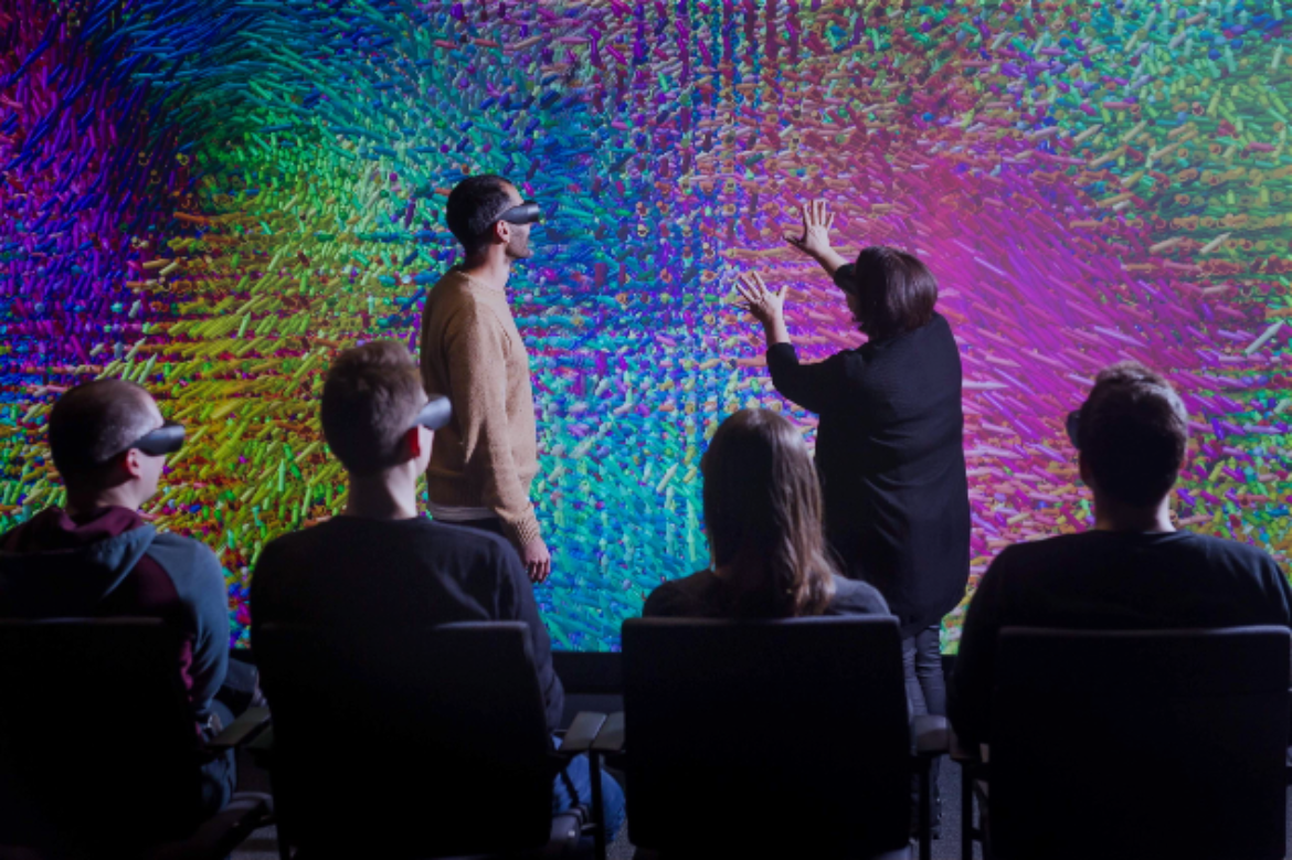 Mehrere Menschen mit VR Brillen sitzen vor einer bunten Wand. Zwei Personen erklären etwas mithilfe von Gesten an dieser Wand.